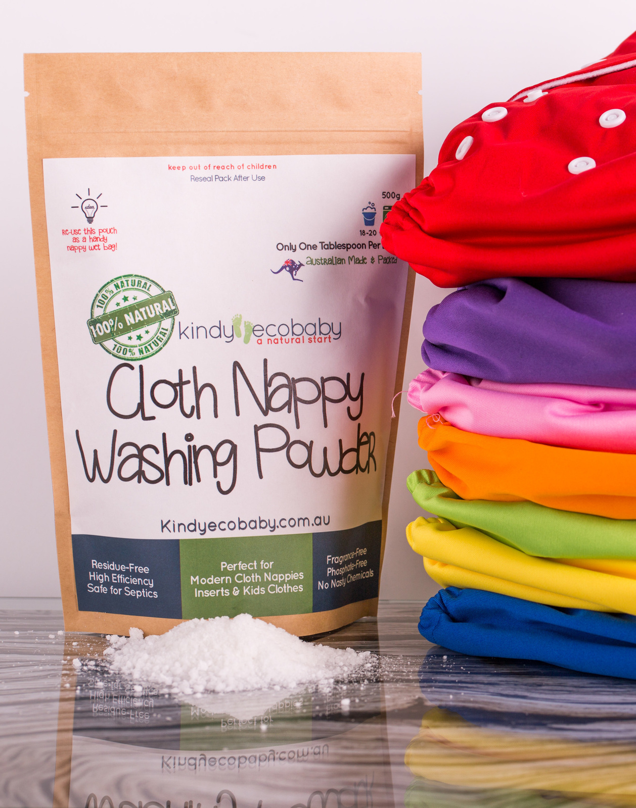 A bag of Kindy Ecobaby Cloth Nappy Washing Powder next to a neat pile of seven modern cloth nappies which are red, purple, orange, pink, yellow, blue and green