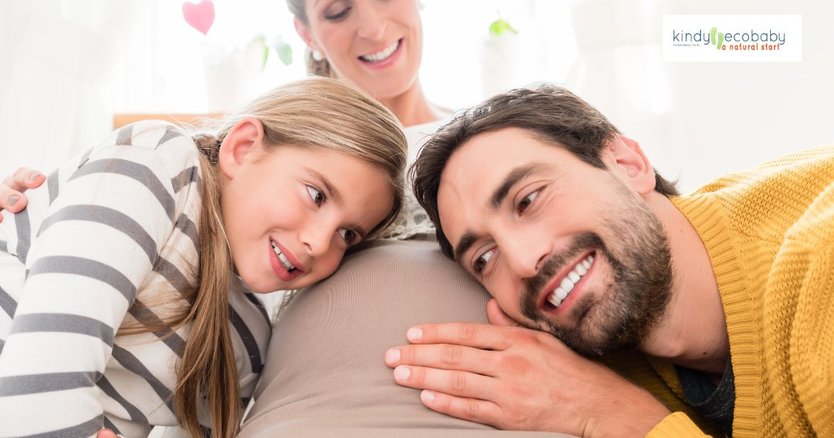 a pregnant person, person and child are smiling while laying on a bed